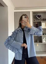 Camisa de corte recto con estampado de cuadros vichy en tonos azul y blanco. Presenta cuello clásico, cierre frontal con botones y volados decorativos que recorren la parte superior de las mangas y los hombros.