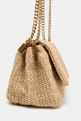 Bolso de hombro formato bandolera con cuerpo en efecto trenzado de color beige. Cuenta con asa de hombro combinada de cadena metálica dorada y tira de cuero sintético marrón, compartimento interior con cremallera y cierre mediante botón imantado.