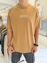 Remera oversize color camel con cuello redondo alto, mangas cortas con costuras raglán decoradas con ribetes blancos y la palabra 'WONDER' bordada en el pecho.