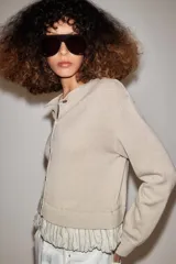 Chaqueta de punto color beige con cuello redondo, manga larga y cierre frontal con botones. Presenta un diseño combinado con volados de tela en el bajo.