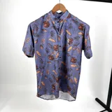 Camisa de manga corta con cuello mao, estampada con hojas en tonos azul y marrón. Presenta un pequeño logo bordado en el pecho.