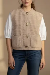 Chaleco tejido color beige con cuello redondo, sin mangas, con cierre frontal de cuatro botones dorados y dos bolsillos frontales.