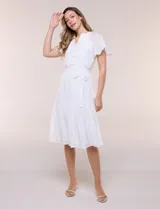 Vestido midi blanco de calce suelto y caída fluida, con escote en V, cuello a la base y apertura abotonada con detalle de puntilla. Cuenta con cintura elastizada con lazo para anudar a tono, mangas cortas con volados y falda con volantes escalonados.