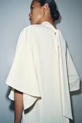 Vestido midi fluido de cuello alto y manga corta tipo capa. Presenta un diseño minimalista con bajo recto y aberturas laterales, con cierre en la espalda.