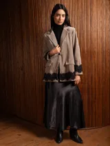 Blazer de corte clásico color beige, con solapa, hombreras y detalle de encaje negro en los puños y el ruedo. Presenta falsos bolsillos frontales y una textura similar al terciopelo.