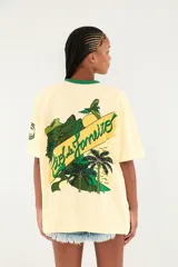 Camiseta Ipanema de manga corta en color amarillo pastel, con cuello redondo en contraste de color verde oscuro. Presenta un pequeño estampado en el pecho con diseño de escudo y palmeras.