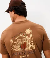 Remera marrón de manga corta con cuello redondo y corte comfort. Presenta una gran estampa en la espalda con diseño de paisaje playero, tablas de surf y tipografía, y estampa en el frente.