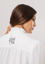 Camisa blanca de cuello redondo con frunces, abotonada y de manga larga con puño abierto.