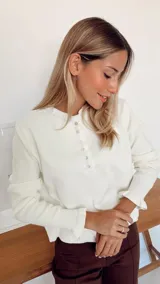 Sweater de punto color crema, con cuello redondo y cierre frontal parcial con botones. Presenta mangas largas con puños acanalados y detalle de volados en los extremos.