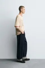 Camiseta de punto tejida color beige claro, corte boxy fit, con cuello redondo y mangas cortas. Presenta estampado desgastado con las letras "LA" en el frente y ribetes en color terracota en cuello y mangas. El bajo es irregular.