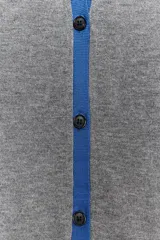 Chaqueta de punto gris de manga larga, cuello redondo y cierre frontal con botones oscuros. Presenta ribetes y detalles en color azul eléctrico en el frente, puños y ruedo.