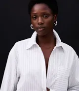 Camisa de mujer de corte maxi oversized, confeccionada en tricolina de algodón certificado. Presenta rayas verticales de hilo teñido, cuello con corbata (gravata), botones frontales, bolsillo en el pecho y puños largos.