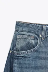 Jeans de tiro alto, color azul medio lavado, con un corte extremadamente ancho y voluminoso (balloon/oversize). Presenta costuras torcidas y paneles de denim de tono ligeramente diferente en los laterales, creando un efecto visual dramático.