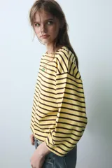 Camiseta oversize de manga larga con diseño de rayas horizontales amarillas y negras. Presenta cuello redondo y un detalle de texto bordado en el centro del pecho.