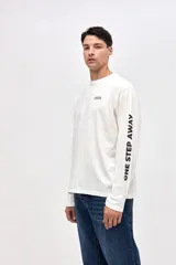 Remera de manga larga color blanco, con cuello redondo y calce clásico. Presenta un pequeño estampado en el pecho y la frase 'ONE STEP AWAY' estampada en una de las mangas.