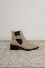 Botineta de cuero charol negro con acabado brillante, diseño de punta fina y taco cuadrado. Presenta un panel elástico lateral y una correa decorativa con hebilla metálica plateada.