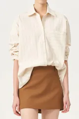 Camisa oversize de color blanco roto con finas rayas verticales en color gris o beige. Posee cuello clásico, manga larga y un bolsillo de parche en el pecho.