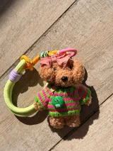 Charm de peluche con forma de osito de color marrón claro, vestido con un sweater tejido a rayas verde lima y fucsia. Lleva un lazo rosa en la cabeza con una perla y un dije verde con el logo de M&M. Incluye un accesorio de cuerda trenzada multicolor para colgar.
