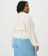 Blusa de viscosa texturada color marfil, con escote en V pronunciado, lazo frontal y corte estilo túnica. Presenta mangas largas abullonadas con puños ajustables mediante tiras y detalles de encaje en el escote, la cintura fruncida y el ruedo.