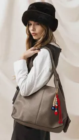 Cartera de símil cuero color marrón topo con diseño de hombro, costuras visibles y cierre superior. Incluye un llavero decorativo con cuentas de madera, dijes de mal de ojo y cordones rojos con un corazón.