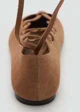 Zapatos chatitos de piel de serraje color marrón, con diseño de tiras cruzadas en el empeine, ojales metálicos dorados y cintas largas para atar alrededor del tobillo. Presentan punta redonda y suela plana.