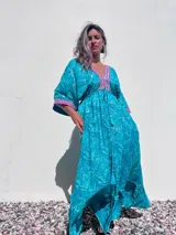 Túnica larga de estilo boho, color turquesa con estampado paisley en tonos azules y dorados. Presenta escote en V profundo con bordados decorativos en color fucsia y blanco, mangas amplias tipo kimono y ajuste en la cintura con cordón. El diseño incluye un ruedo con volado y terminaciones con borlas.