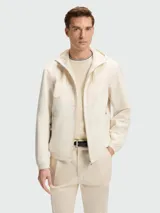 Chaqueta ligera con capucha, cierre frontal y puños elásticos, color beige claro. Presenta un diseño con costuras en forma de chevron en el pecho.