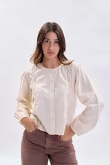 Blusa casual de color crudo con mangas largas abullonadas, cuello redondo y cierre frontal con botones. Presenta delicados detalles de encaje y pliegues en la parte frontal.