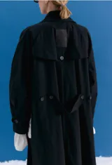 Trench midi de color negro, confeccionado en una mezcla de algodón y nylon con propiedades repelentes al agua. Presenta un diseño holgado con tajos laterales, bolsillos internos y detalle de etiqueta en la espalda.