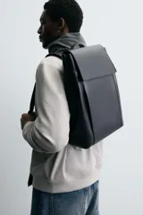 Mochila negra de poliuretano, con solapa y cierre de hebilla. Tiene un compartimento principal con espacio para dispositivos electrónicos de hasta 13 pulgadas y un bolsillo con cierre. En la parte frontal tiene un bolsillo con cierre y bolsillos planos en la parte frontal y posterior. Tiene asa de mano y dos asas de hombro ajustables y acolchadas.