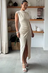 Vestido largo sin mangas de color beige, con cuello redondo y diseño drapeado en la cintura que crea un efecto fruncido. Presenta una abertura lateral pronunciada en la falda que aporta movimiento y un toque sofisticado.