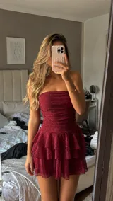 Vestido corto strapless de color bordó con acabado brillante. Presenta un diseño ajustado en el torso con frunces y una falda con volados superpuestos.