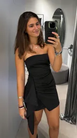 Vestido corto strapless de color negro con sutiles destellos brillantes. Presenta un diseño ajustado con drapeado lateral y un detalle de solapa asimétrica que cae sobre la falda.