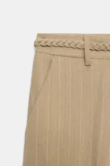 Pantalón de tiro medio color beige con sutiles rayas verticales blancas, de corte recto y amplio. Cuenta con bolsillos delanteros y un cinturón trenzado del mismo tejido con cordón ajustable.