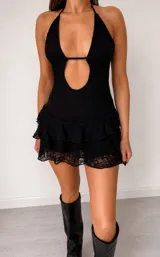 Vestido corto de encaje negro con diseño halter, escote profundo y abertura frontal. Presenta una falda con volados superpuestos y terminación en encaje transparente.