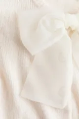 Saco de punto suave color crema, de manga corta y cuello redondo. Presenta cierre frontal con botones forrados y una moña decorativa de organza transparente en el escote. Posee terminaciones acanaladas en puños y ruedo.