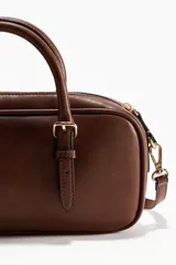 Morral rectangular de cuero sintético marrón, con cierre superior, asas cortas y correas con detalles de hebillas metálicas.