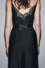 Cinturón tipo fajín ancho de piel sintética negra, con forma curva en la parte superior e inferior. Presenta detalles metálicos dorados en los laterales y se ajusta mediante lazos largos del mismo material.