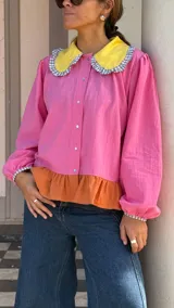 Camisa de manga larga en color rosa, con cuello Peter Pan en color amarillo y detalle de volados en los bordes. Presenta cierre frontal con botones y puños elásticos con detalle de volados.