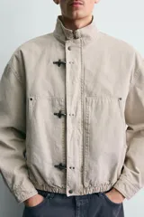 Cazadora relaxed fit confeccionada en tejido de algodón de color beige, con cuello subido y manga larga. Cuenta con bolsillos de plastrón en la cadera, bajo elástico y cierre frontal con cremallera oculta y alamares metálicos.