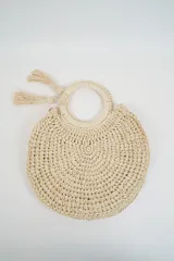 Cartera redonda tipo bolso de mano, tejida en crochet de color beige, con asas circulares rígidas y borlas colgantes decorativas.