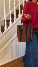 Matera de cuero con diseño de paneles verticales en combinación de colores azul, verde, marrón y bordó. Cuenta con dos asas cortas de cuero y estructura rígida.