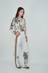 Pantalón de lino color celeste claro, de corte holgado y pierna recta. Presenta una franja lateral decorativa con estampado geométrico y detalles de palmeras estampadas en la parte inferior de las piernas.