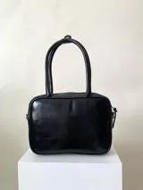 Cartera rígida negra con detalles en rojo y tachas plateadas. Tiene herrajes y cierre metálico plateado marca YKK, asa fina para llevar al hombro o en la mano y correa larga extra regulable. El interior es forrado, tiene un bolsillo con cierre enmarcado en cuero, y un segundo bolsillo plaqué.