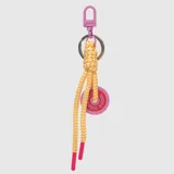 Charm para bolso con mosquetón metálico rosado, argolla plateada y cordón trenzado en tonos amarillo y blanco con punteras fucsias. Incluye un dije circular de silicona fucsia con el logo de la marca grabado.