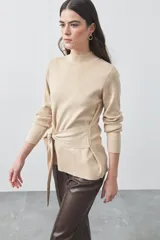 Polera de tejido de punto color beige, con cuello alto, manga larga y detalle de lazo ajustable en la cintura que aporta estructura a la silueta.