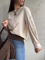 Blusa de color beige con diseño de corte holgado y mangas largas abullonadas. Presenta detalles bordados en tono sobre tono en las mangas y puños ajustados.