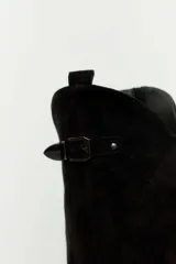 Bota de caña alta estilo slouchy confeccionada en gamuza sintética de color negro, con punta fina y taco bajo. Presenta solapa superior plegada con apliques de tachas metálicas plateadas y una tira con hebilla decorativa en la parte superior de la caña.