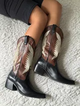Botas texanas de caña media confeccionadas en cuero, con diseño bicolor que combina negro en la base y puntera, y marrón con detalles en cuero texturizado brillante en la caña. Presentan un taco cuadrado de 5 cm y diseño sin cierre.