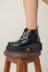 Bota corta de cuero negro con suela track dentada y taco cuadrado. Presenta una tira decorativa en el empeine con una hebilla metálica plateada y tirador en la parte trasera.